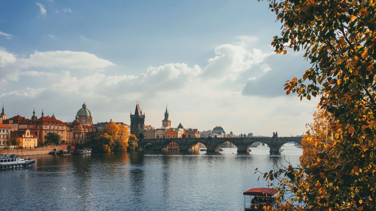 A scenic landscape of Prague&apos;s Charles Bridge during autumn, with golden leaves, vibrant street performers, and a warm sunset casting a glow on the Vltava River